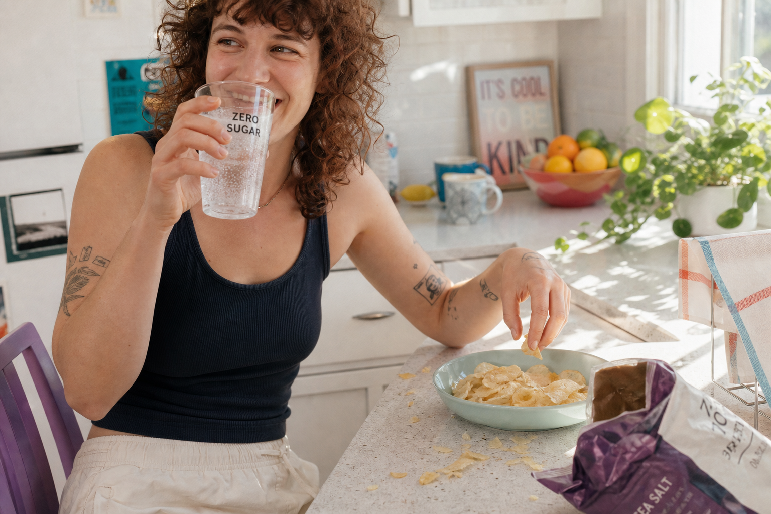 Vrouw met krullend haar drinkt uit een glas met 'zero sugar' in een zonnige keuken naast een kom chips.